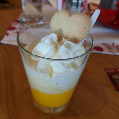A wedge-shaped glass filled with a bright yellow liquid, topped with a swirl of squirty cream and a heart-shaped biscuit.