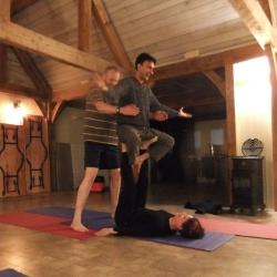 Three people engaged in acro yoga.  One is laying on the floor with their feet in the air.  A second is balanced cross-legged on their feet.  The third is standing behind them, providing support.