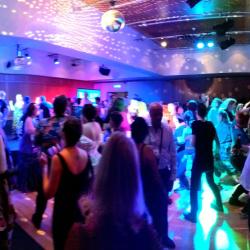 A crowd dance under multicoloured lights, in a dark room.