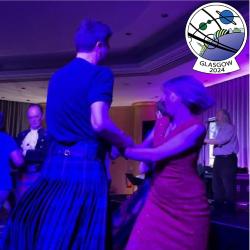 Two people dancing.  A man in a kilt and a woman in a pink dress hold hands and swing around each other.  In the corner of the image is the Glasgow in 2024 logo.